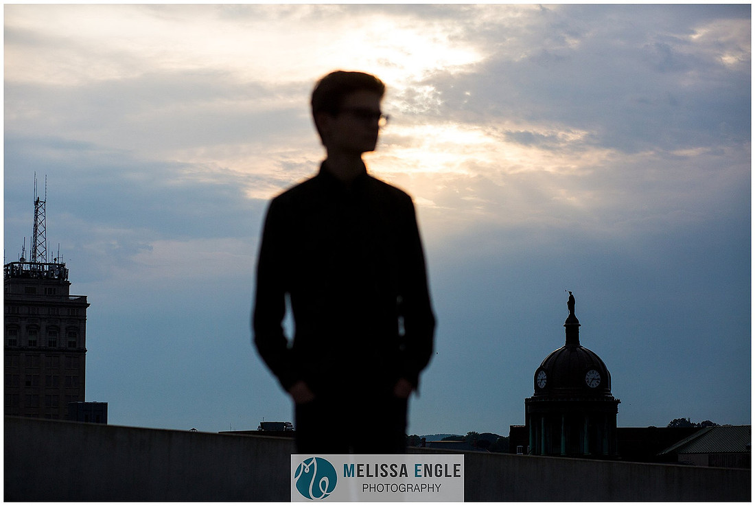 Creative senior portrait of a guy wearing glasses silhouetted against a blue sunset sky in an urban setting in downtown Lancaster City.