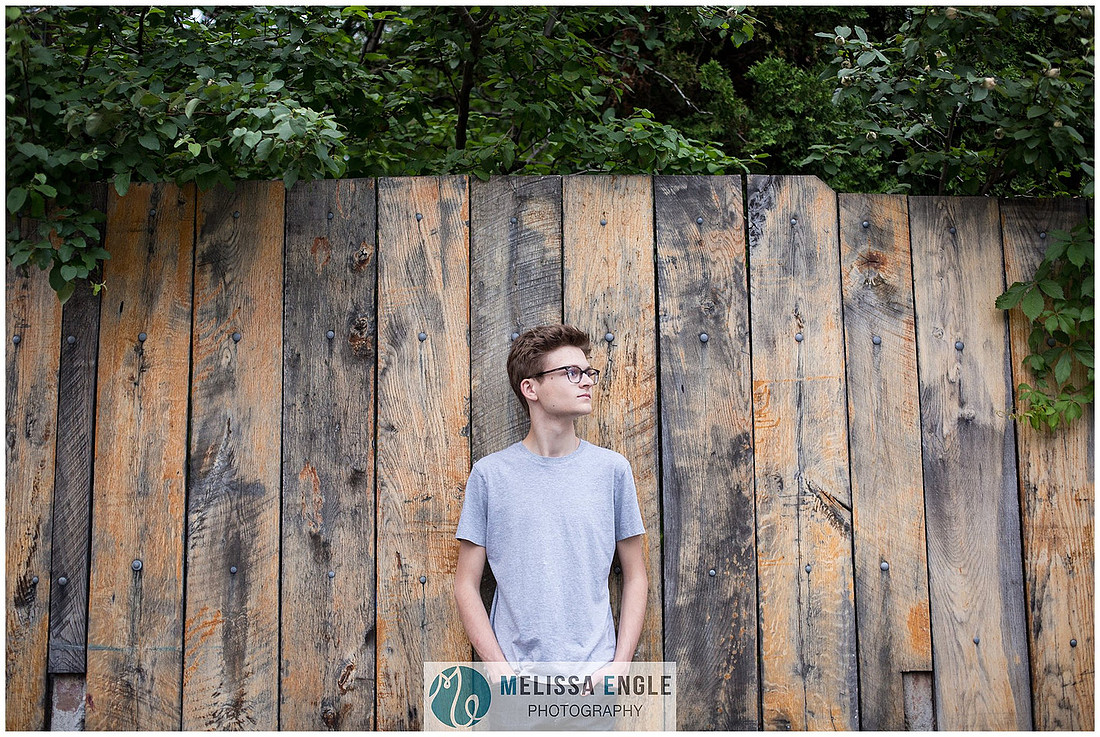 Creative senior portrait of a young hip guy wearing glasses and a grey shirt against a wooden fence in downtown Lancaster City.