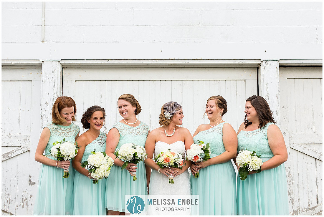 Photo of a bride and her bridesmaids wearing mint green dresses in front of a white barn laughing.