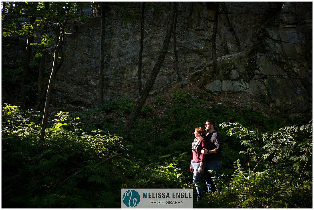 Engagement photo of a couple in the middle of the woods surrounded by trees, rocks and sunlight at Money Rocks County Park.