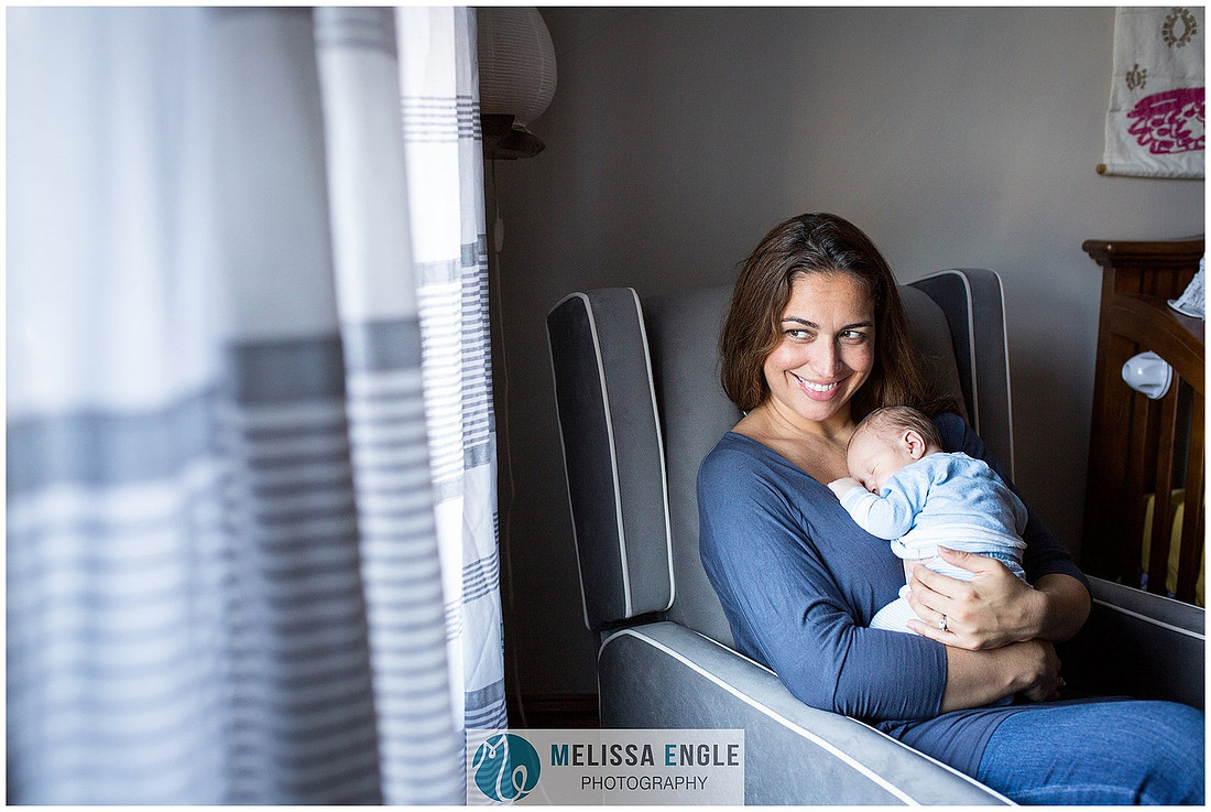 Happy young mother sits next to a window while holding her newborn baby boy.