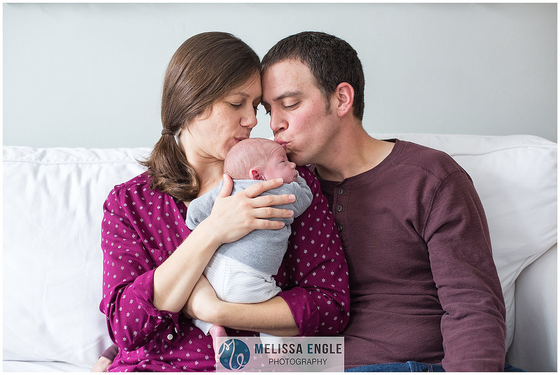 Photo of a young couple sitting on a white couch kissing their newborn baby boy.