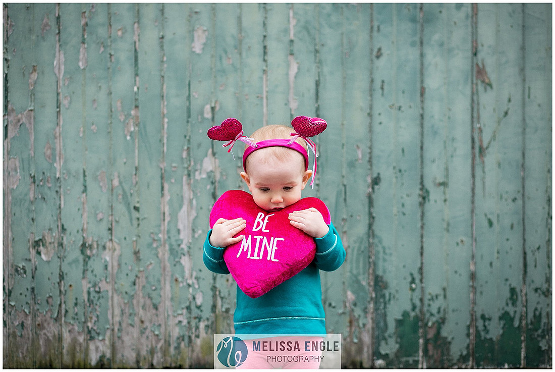 Photo of a toddler wearing a heart antennae headband holding a bright pink pillow that says "be mine".